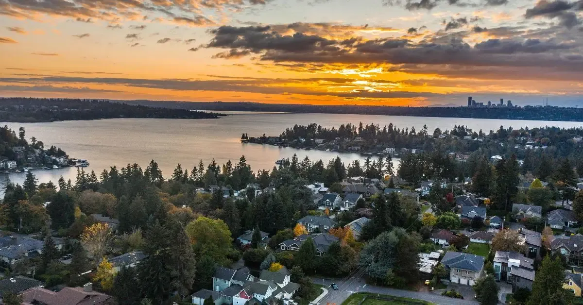 Aerial view of a tree-lined lakeside neighborhood at sunset, with a city skyline on the far shore