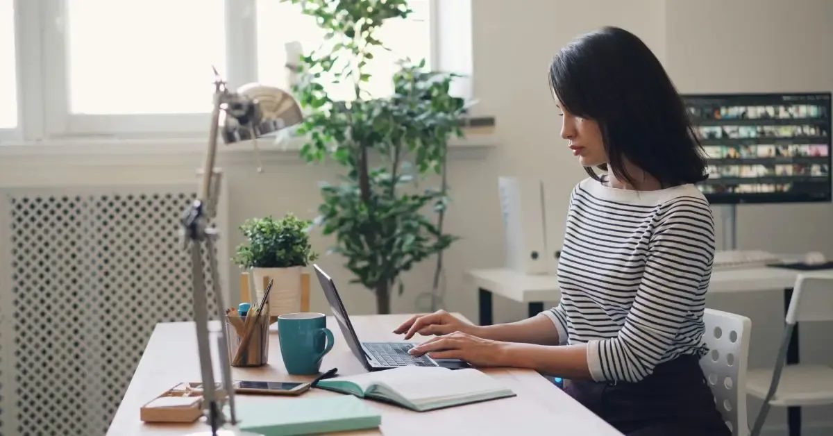 A woman with dark hair typing on a laptop at a wooden desk, beside a mug, a notebook and a potted plant
