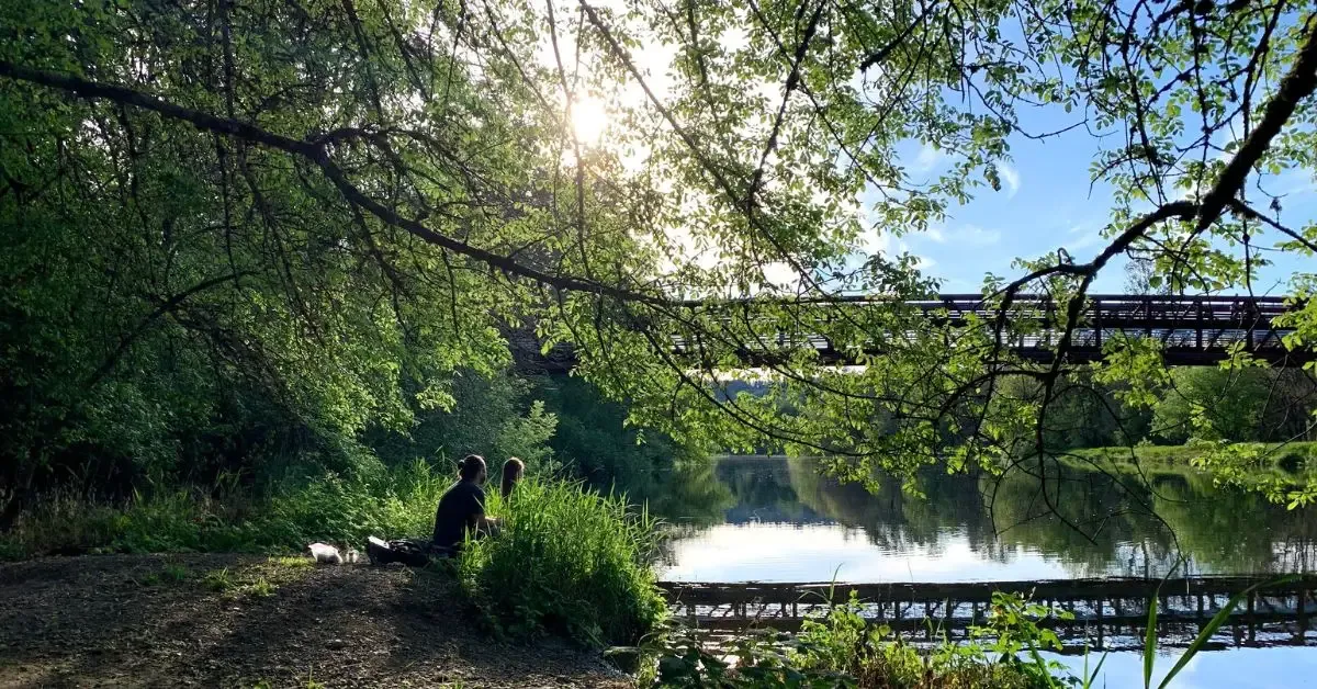 Two people sitting on a grassy riverbank under leafy trees, with a bridge crossing the river behind them