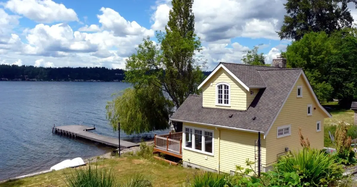 A yellow two-story lakeside cottage with a wooden deck and a dock, on a calm lake under a partly cloudy sky