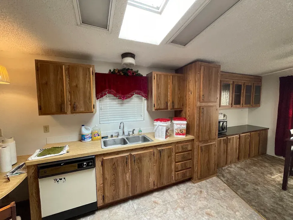 The kitchen of an Auburn, WA manufactured home, with dark wood-grain cabinets, laminate counters, an older dishwasher, a textured ceiling and a skylight