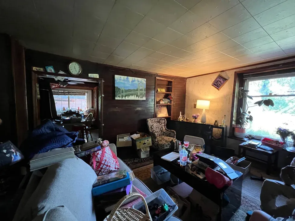 The living room of a Vancouver, WA house, with dark wood-panelled walls, a tiled ceiling, and a sofa and coffee table piled with books, boxes and belongings